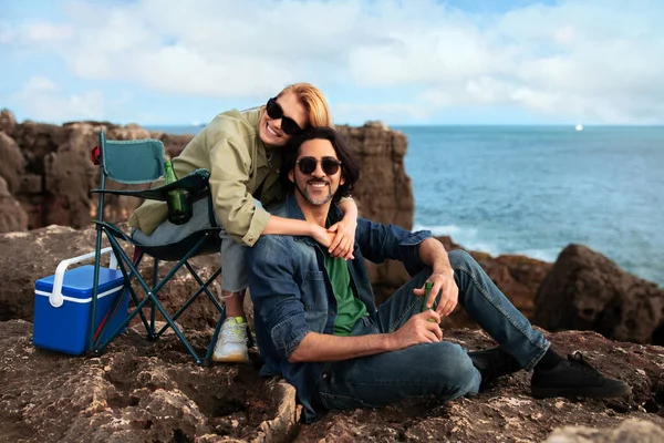 千年快乐伴侣在靠近海洋的海滩岩石上露营椅中放松身心 在自然景观中微笑男人和女人 喝汽水和拥抱 享受户外休闲 复制空间 — 图库照片