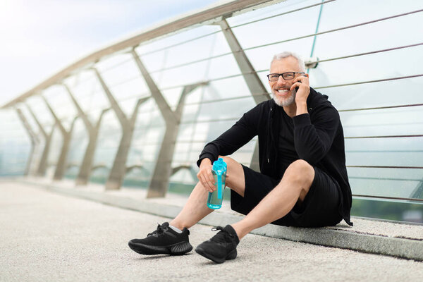 Positive well-fit senior sportsman in black sportswear sitting on bridge, holding bottle with water, have phone conversation while resting after workout outdoor, looking at copy space