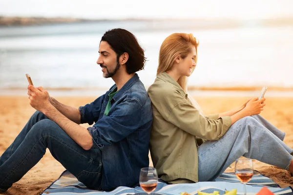 ビーチの屋外でピクニックをしながら スマートフォンでリラックスした幸せな若いカップル 笑顔の男と女が海の近くで再生され 携帯電話を使用して後ろに座って 無料のスペースでサイドビュー — ストック写真
