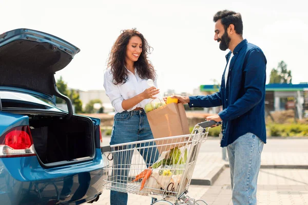 Married middle eastern couple putting paper bag full of healthy food into car, man helping wife, standing outdoors on parking near shopping mall