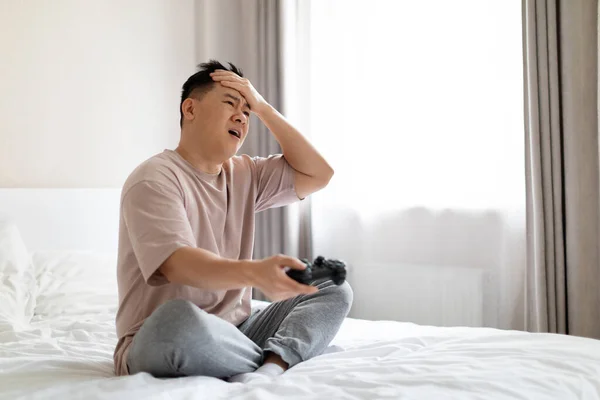 电子游戏成瘾的概念 穿着睡衣坐在床上 拿着控制杆 摸着头 在家里玩电子游戏 复制空间的不快乐的亚洲中年男人失败者 — 图库照片