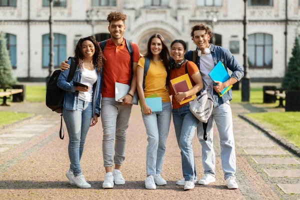 快乐的国际学生站在大学大楼近旁 快乐的年轻人微笑拥抱 多元文化的朋友背着背包和工作手册站在外面的画像 — 图库照片