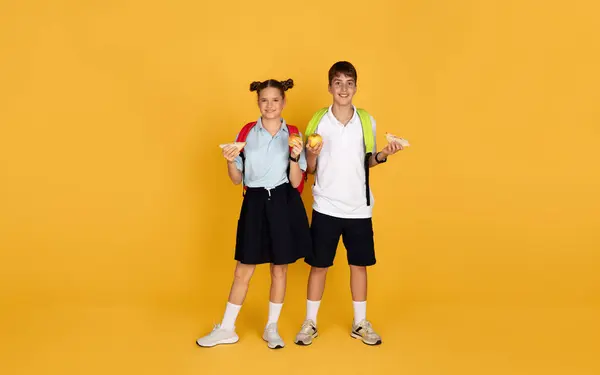 バックパック付きの笑顔10代のヨーロッパの男の子と女の子は 黄色のスタジオの背景に 完全な長さに隔離されたリンゴとサンドイッチを保持します 学校での勉強 食べ物や友情のための休憩 — ストック写真