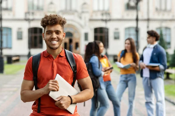 High Education Concept. Portrait of happy black male student smiling at camera while posing outdoors in campus, his classmates and university building on background, selective focus