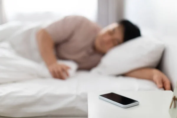 技術と人々の概念 現代のスマートフォンに焦点を当てます黒の空の画面上のベッドサイドテーブルアジアの男の近くのベッドで寝て自宅で朝 ぼやけた背景 — ストック写真
