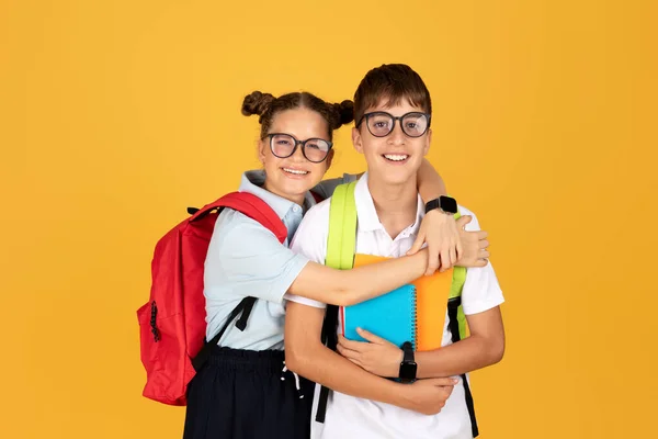 黄色のスタジオの背景に隔離されたノートブック付きのバックパック抱擁の男の子とメガネで幸せなティーンヨーロッパの女の子 子供時代 学校での勉強 — ストック写真