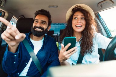 Gülümseyen orta doğulu çift lüks arabanın içinde oturuyor, direksiyon tutan heyecanlı kadın, cep telefonlu adam yolu işaret ediyor, dijital harita uygulaması kullanıyor.