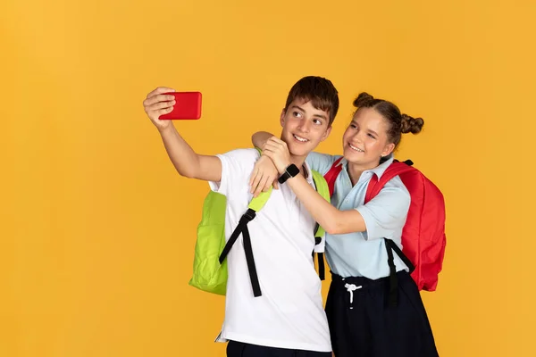 快乐的少年欧洲男孩和女孩背着背包拥抱 在智能手机上自拍 在黄色的工作室背景下被隔离 学校教育应用 照片和装置 — 图库照片