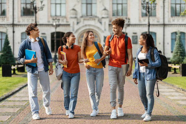 Happy university students walking together on campus, gropu of cheerful young multiethnic people chatting and laughing outdoors during break, carrying backpacks and workbooks, full length shot