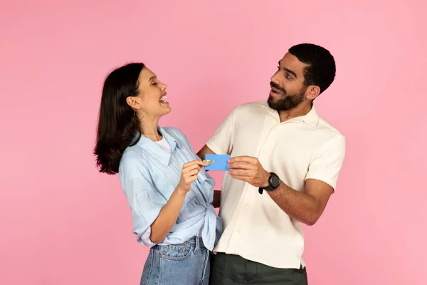 兴奋的千禧年快乐的他的惊慌失措的男人和女人拿着蓝色塑料信用卡 面带微笑地看着对方 被粉红色的背景隔离了 — 图库照片