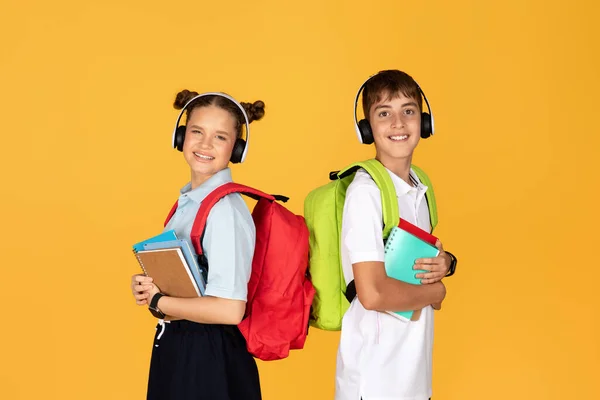 快乐的欧洲女孩背着背包 男孩戴着耳机 带着笔记本 与黄色工作室背景隔离 听音乐 友谊和童年 — 图库照片