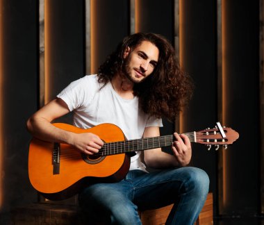 Yakışıklı İspanyol akustik gitarlı genç adam stüdyoda siyah şarkısını çalarken kameraya bakıyor. Müzikal Enstrümanla Oturan Profesyonel Müzisyen