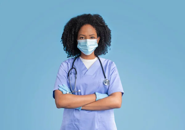严重自信的年轻黑人女医生 身穿蓝色制服 戴着防护面具和手套 双手交叉放在胸前 与蓝色工作室背景隔离 — 图库照片