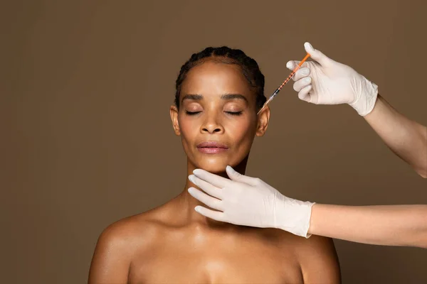 Cosmetic beauty injections concept. Attractive black middle aged woman getting facial injection in cheekbones zone, standing over brown studio background, free space