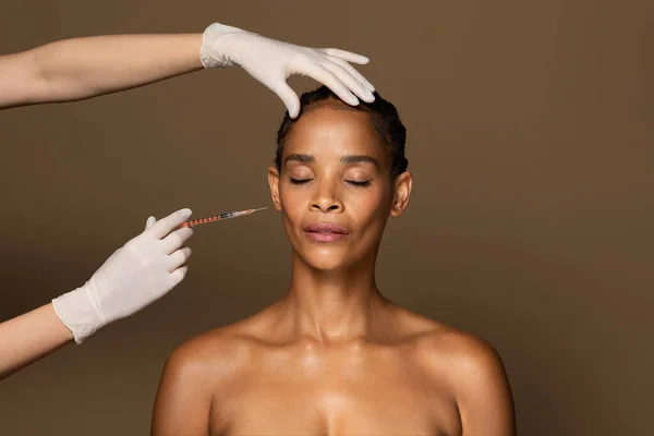 美丽的非洲裔美国中年妇女在颧骨区接受面部注射 站在棕色工作室背景之上 化妆品注射概念 — 图库照片