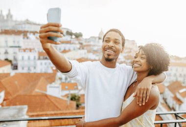 Neşeli, Afro-Amerikalı, giyinmiş bayanı kucaklayan, selfie çeken, sosyal ağlar için fotoğraf çeken, randevunun keyfini çıkaran, seyahat eden, açık havada gezen genç bir adam. Blog, video çağrısı, aşk ilişkisi, tatiller, tatil ve romantizm