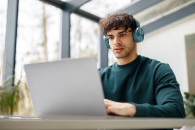E-öğrenme konsepti. Kablosuz kulaklıklı Avrupalı adam dizüstü bilgisayara bakıyor, çevrimiçi eğitim, ders ya da sınıfta oturuyor ya da kütüphanede oturuyor.
