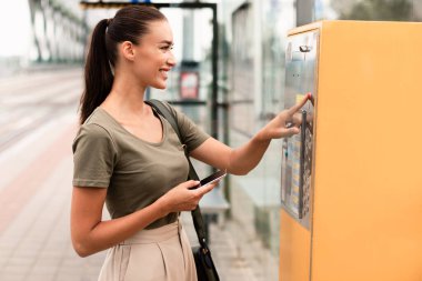 Modern Tramvay Durağı 'nda bilet makinesini kullanan, akıllı telefon tutan ve düğmeye basan, toplu taşıma için kontaksız ödeme yapan mutlu yolcu kadınlar. Yan Görünüm Görüntüsü