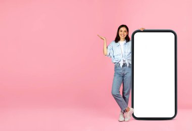 Neşeli, güzel, günlük kıyafetler giyen, beyaz ekranlı, hareketli, pembe arkaplanlı, büyük akıllı telefonlu bir poz veren güzel bir kadın. Güzel online anlaşma, teklif