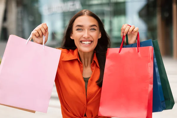 季节性销售 微笑的女人手握彩纸购物袋站在市区的广告商摊位上 凝视着相机 买家夫人在户外展示套件 — 图库照片
