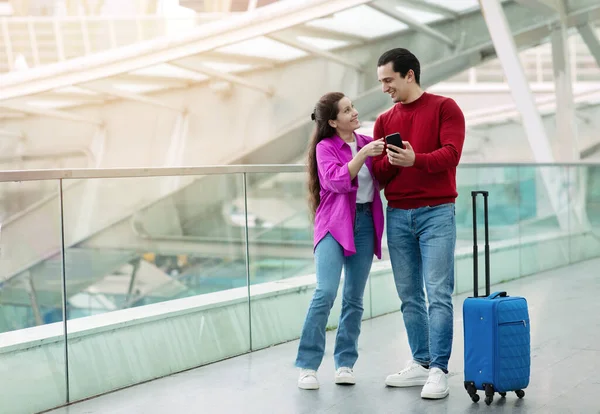 旅行アプリ モダンな空港に立つスマートフォンとスーツケースでポーズをとる幸せな観光客カップル 携帯電話を指している女性お勧めのアプリケーションと予約チケットオンライン コピースペース — ストック写真