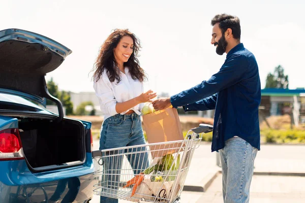 Happy middle eastern couple packing grocery bags into a car trunk, arab man giving paper bag to wife, standing near shopping cart full of fresh food