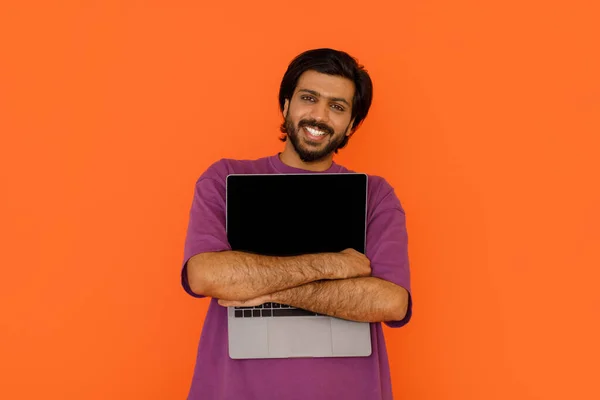 麻薬中毒だ 積極的なハンサムな若いインド人男性抱擁ラップトップで黒の空の画面上のオレンジのスタジオの背景 オンラインオファー アプリケーション ウェブサイトの広告 コピースペースのためのモックアップ — ストック写真