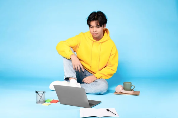 アジアの学生の男は ラップトップコンピュータの学習や宿題をオンラインで行うと 青いスタジオの背景にポーズを床に座っている インターネット教育 近代的な研究と技術の概念 — ストック写真