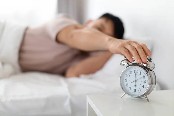 Wake up, good morning. Asian man wearing pajamas turning off ringing alarm clock stand on bedside table, waking up in the morning, selective focus on watch, blurred background