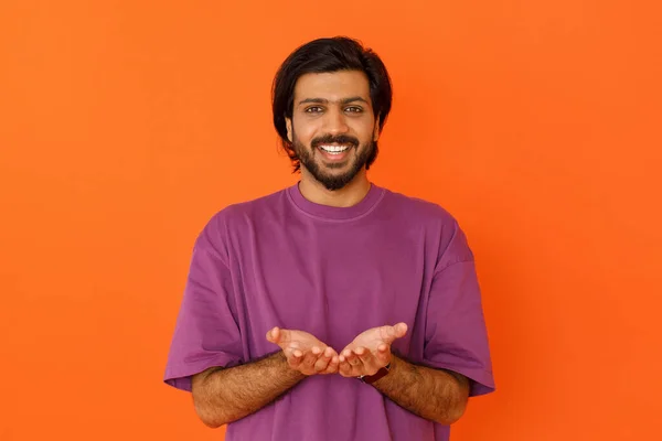 彼の胸の横に開いて手を保持しているカジュアルな紫のTシャツを着て幸せな笑顔若いインド人男性 彼の手のひらに見えない何かを示すHindu男 オレンジのスタジオの背景に孤立 — ストック写真
