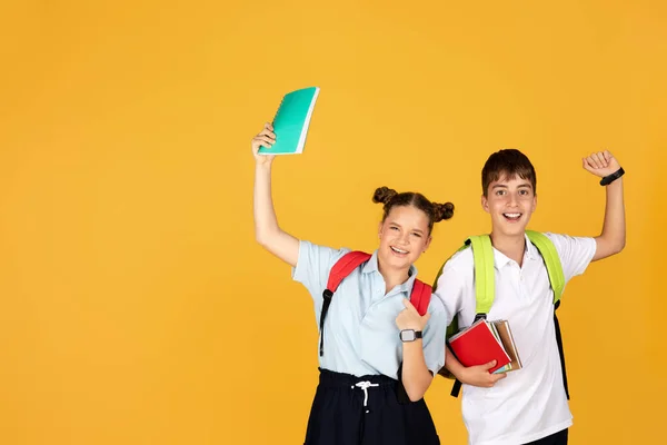 快乐的欧洲女孩背着背包 男孩拿着笔记本庆祝开始学习 被孤立在黄色的工作室背景下 学校教育 — 图库照片