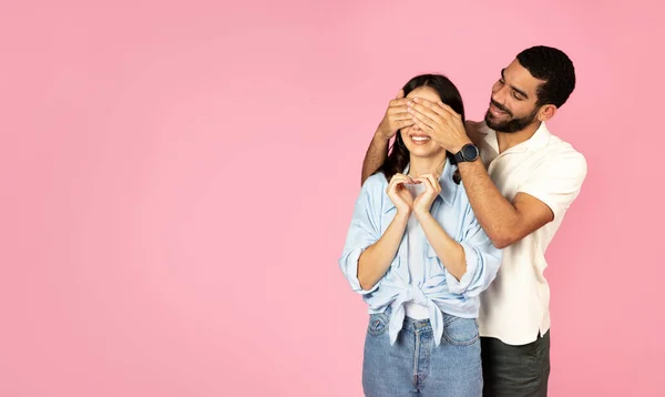 可爱的年轻的中东男子覆盖兴奋的女人他的束腰或妻子的眼睛 孤立在粉红的工作室背景 庆祝结婚一周年的快乐新婚夫妇 复制空间 — 图库照片