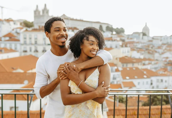 Smiling young african american guy hugging lady in dress, look at copy space in city, enjoy honeymoon, date and travel, outdoor. Romantic trip, love relationships, holiday vacations and spare time