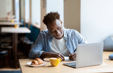 Happy black man studying remotely in front of laptop, taking notes during online class or lecture at city cafe. Industrious African American guy writing in copybook, working on project at coffee shop