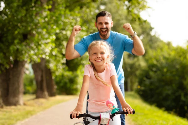幸せな父親は子供の娘に公園で自転車に乗るように教え パパはバックグラウンドで拳を振り 夏の楽しみと屋外を散歩します 笑顔の子供の学習とパパからの助けを借りて自転車に乗る — ストック写真
