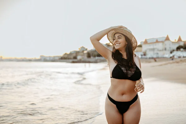 スイムウェアで幸せな若いヨーロッパの女性と日没に沿って海岸線を歩きながらビーチに立っているわら帽子 無料のスペースでポーズして見る — ストック写真