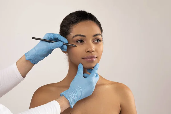 Plastic surgery, injection cosmetology concept. Plastic surgeon hands in medical gloves holding pen, making preparation lines on beautiful young black woman face, grey background, copy space