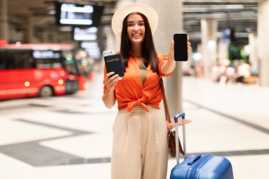 Biletlerinizi internetten ayırtın. Uçuş kartı cep telefonu ekranını gösteren Mutlu Yolcu Kadın, Otobüs Taşıma Teklifi için Mockup Modern İstasyonda Bavulla Bekliyor. Mockup, Seçici Odaklanma