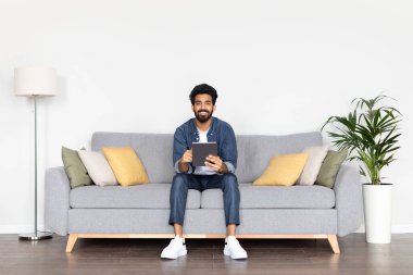 Mutlu, yakışıklı, sakallı, genç bir Arap evdeki dijital tableti kullanarak beyaz arka planda koltukta oturuyor. Dijital göçebe web sörfü, sohbet, alışveriş, online film izleme