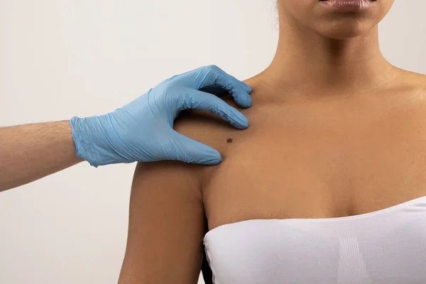 Oncologist wearing blue medical gloves examining patient half-naked black woman skin in clinic, touching mole nevus, isolated on grey background. Cancer awareness, oncology concept