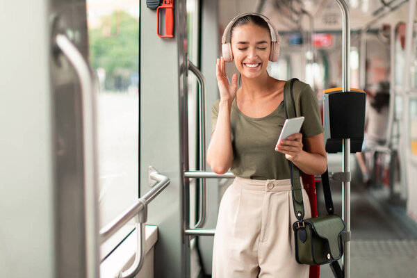 Cheerful Passenger Woman In Modern Tram Holding Phone Listening To Music In Earphones, Commuting And Enjoying Comfort Of City Public Transport Standing Inside. Transportation Leisure Concept