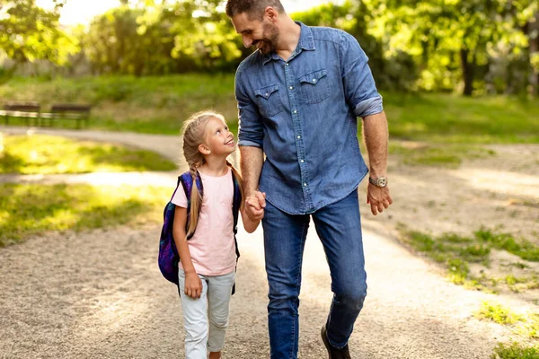 学校初日でした ハッピー パパが小学校1年生を率い 娘と手を握って話し 晴れた日に公園を歩いている — ストック写真