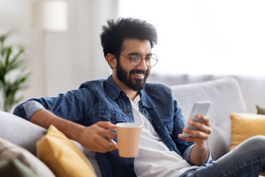 Zeki telefon ve kahveyle koltukta dinlenen gülümseyen Hintli genç bir adamın portresi, online haberleri okuyan neşeli bir doğulu çocuk veya evdeki cep telefonuyla sosyal ağları tarayan