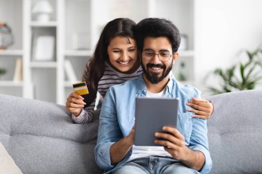 Neşeli, mutlu, genç, doğulu çift oturma odasında koltukta oturuyor, dijital tablet ve sarı plastik kredi kartı kullanıyorlar, evden alışveriş yapıyorlar veya bankacılık yapıyorlar.
