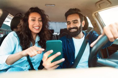 Mutlu Arap adam sürücüsü, güzel kız arkadaşıyla yeni arabalarıyla seyahat ediyor, cep telefonuna GPS takıyor, akıllı telefon ekranına bakan çift, gösterge panelinden vurulmuş.