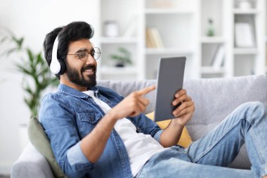 Evde dijital tabletle dinlenen kablosuz kulaklık takan gülümseyen Hintli adam, oturma odasında modern Gadget ile dinlenen yakışıklı genç doğu çocuğu, sosyal ağları tarayan