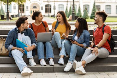 Üniversite yaşam tarzı. Bir grup öğrenci dışarıda birlikte dinleniyor, kampüs parkında merdivenlerde oturuyor, neşeli erkek ve kadınlar sohbet ediyor ve gülüyor, derslerden sonra çok ırklı arkadaşlar dinleniyor.