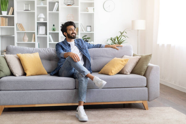 Расслабься Улыбающийся молодой индиец сидит на куше и смотрит в сторону, счастливый симпатичный расслабленный восточный парень отдыхает в гостиной, наслаждаясь домашним комфортом и выходные отдыха, копировать пространство
