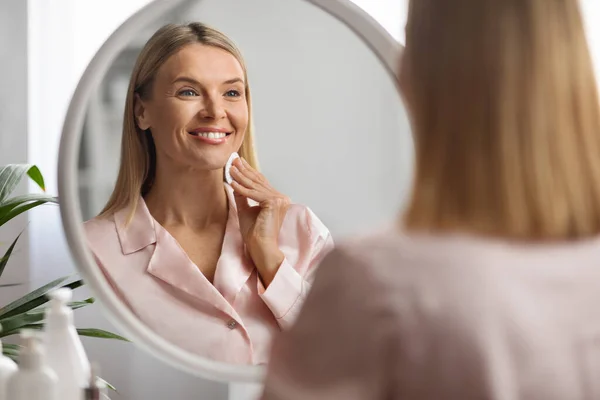 成熟したスキンルーチン 笑顔の中年女性 綿パッドで顔をクレンジングし 鏡で見る 美しい女性は 家庭で美容ケア治療を行う 反射に選択的な焦点 — ストック写真