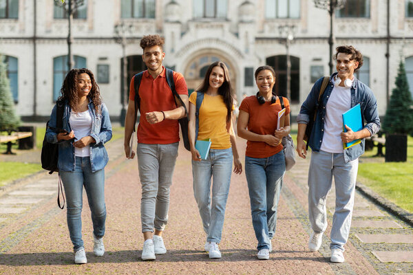 Group of cheerful college students walking out of campus together, happy smiling multiethnic young people with backpacks and workbooks posing at camera outdoors, having break in classes, full length